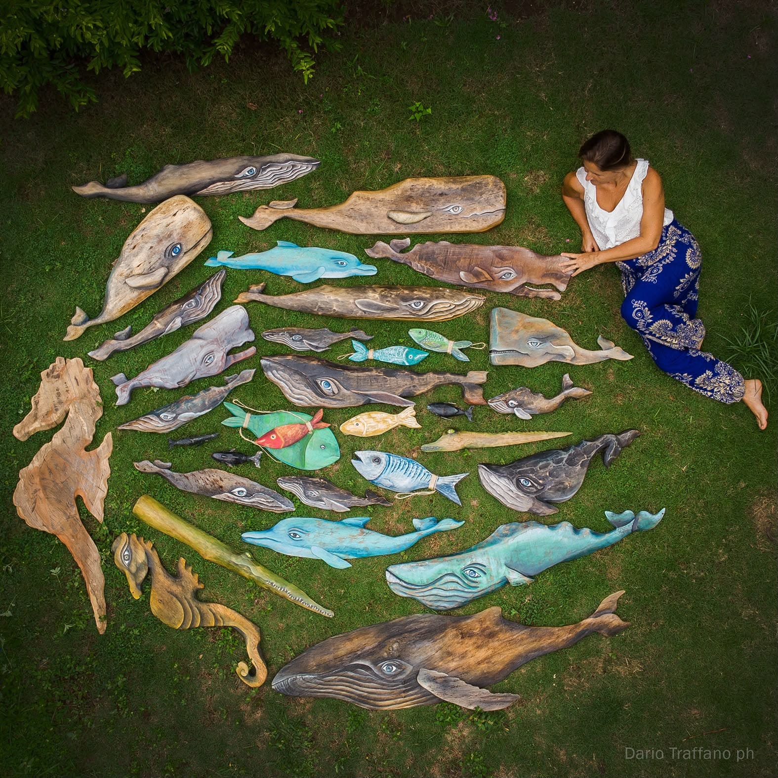 Una artista sentada en el césped junto a una colección de ballenas y peces tallados y pintados en madera